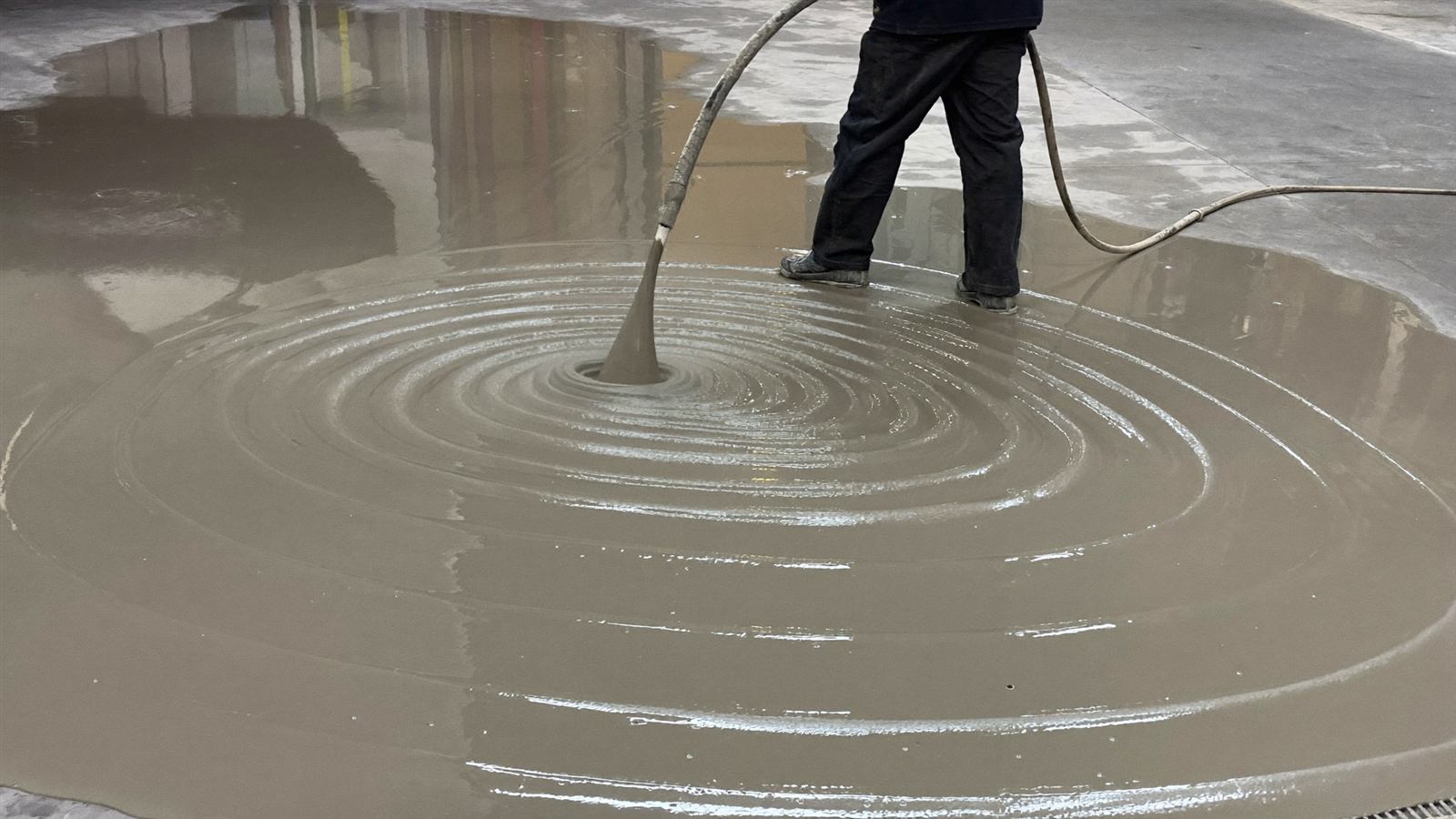 Selbstverlaufender Zementfließestrich CemFlow wird über einen Schlauch in einer Industriehalle verpumpt, reflektierende nasse Oberfläche